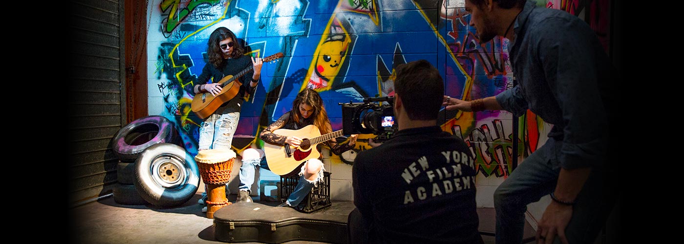 Two NYFA Australia students are filmed playing guitar near a graffitied wall