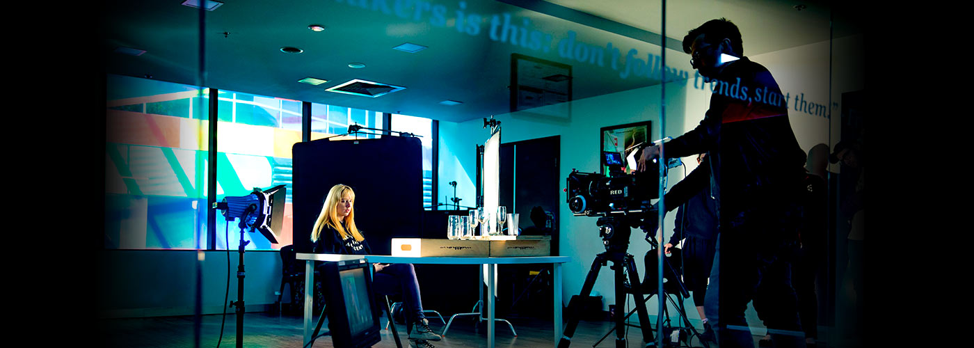 A NYFA Student points a camera at a blonde woman sitting at a desk in a NYFA classroom.
